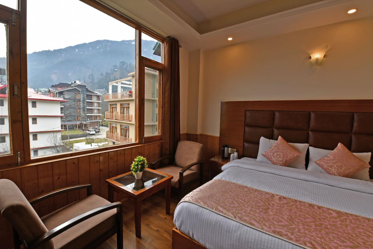 A hotel room with a large bed, a wooden headboard, a small table with a plant, a chair, and a view of the mountains through the window.