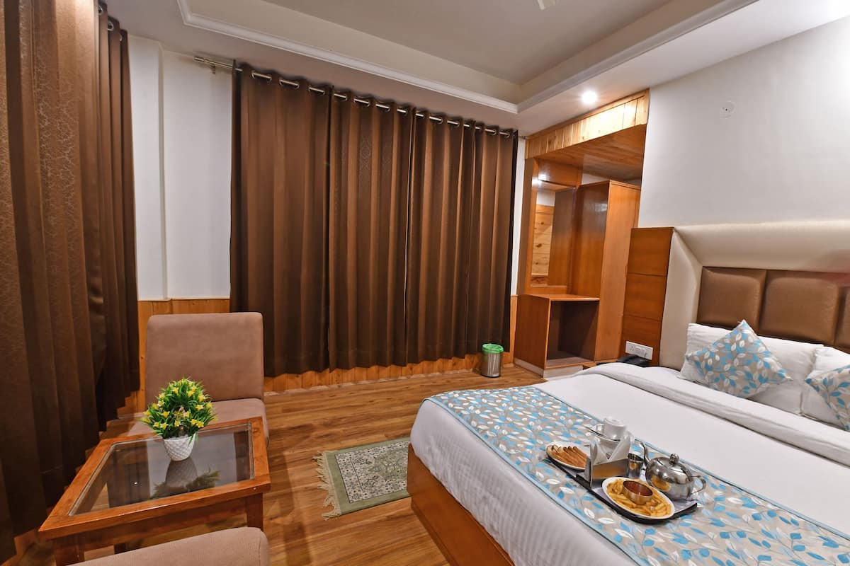 A hotel room with a bed, a small table with a plant, a chair, a wooden floor, a large window with curtains, and a wardrobe.