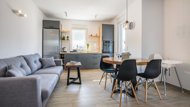 A modern living room with a grey sofa, a dining table with chairs, and a kitchen area with a refrigerator, oven, and window. - Apartments Pejakovic (Vir)