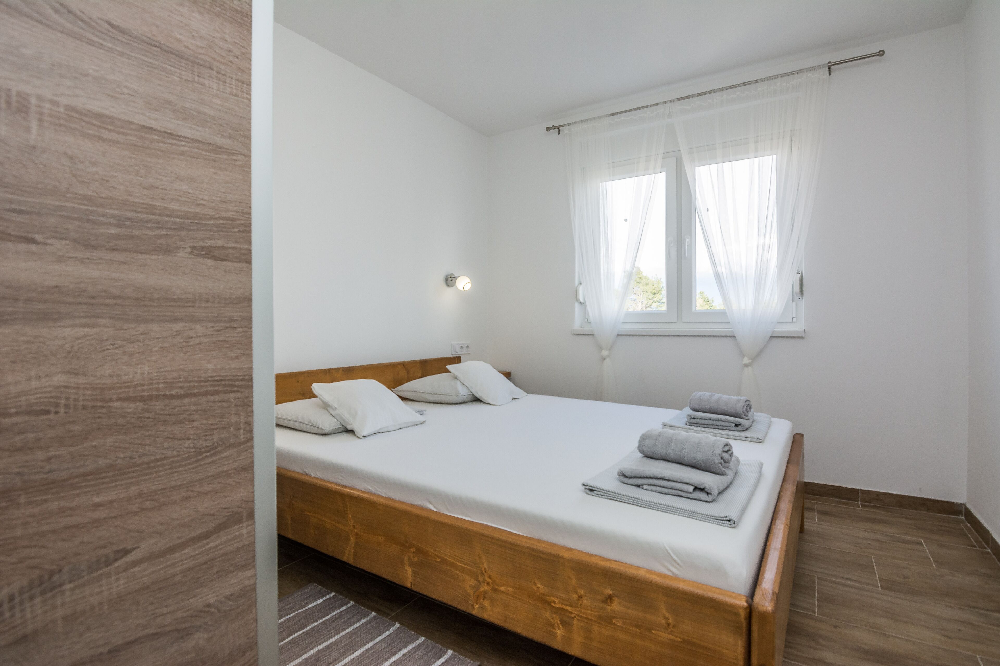 Habitación individual con cama de madera, ropa de cama blanca, ventana con cortinas translúcidas y lámpara de noche.