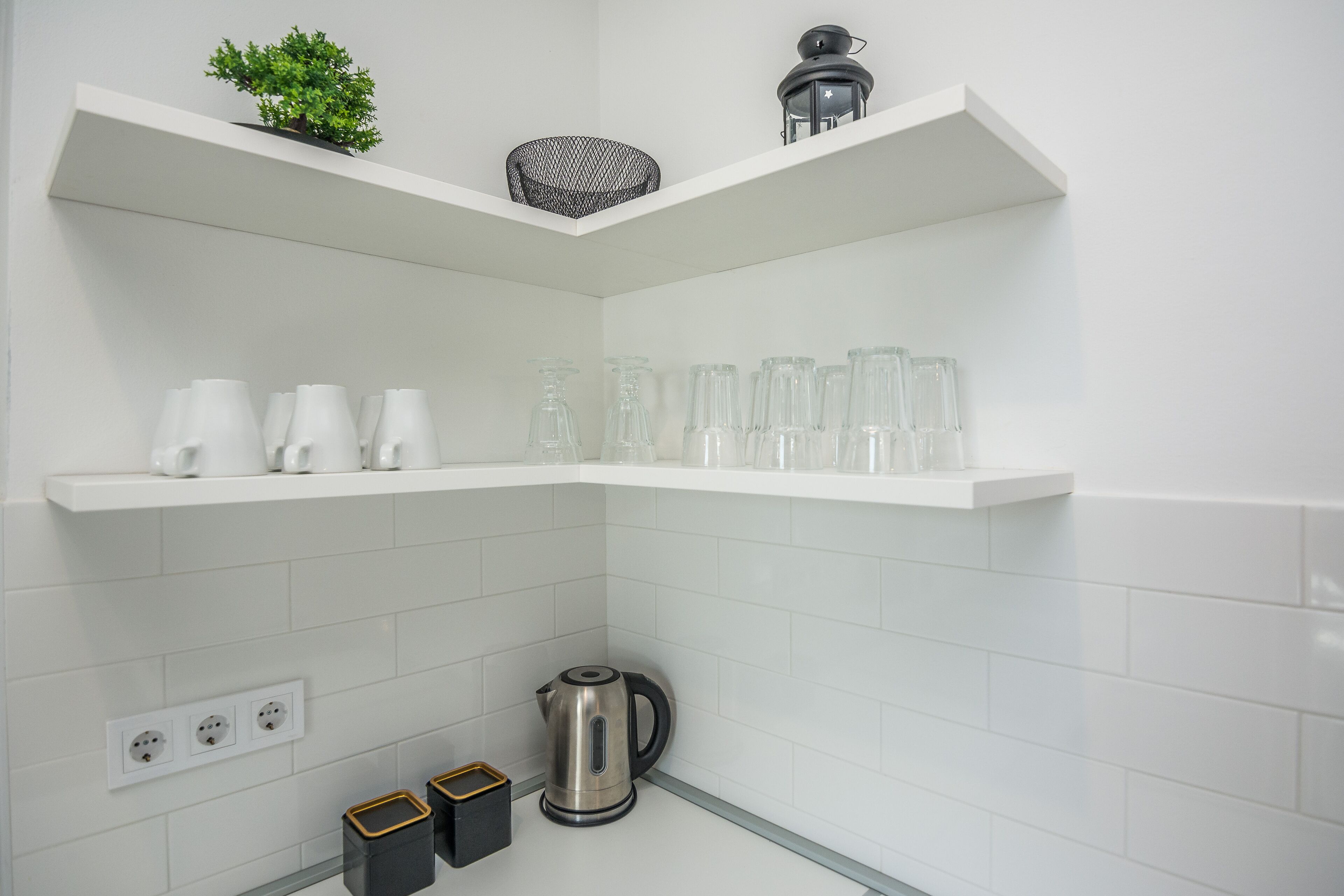 A modern kitchen with white tiled walls, two white shelves, a small potted plant, a black lantern, and a kettle.