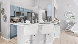 A modern kitchen with blue cabinets, a white island with bar stools, stainless steel appliances, and decorative items. - ⭐Modern & Luxurious 4 Bed Home | Gated Resort | SOUTH Facing Pool | Mins to WDW⭐ (Davenport)