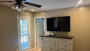 Ein Zimmer mit Deckenventilator, einem großen Flachbildfernseher an der Wand, einer weißen Kommode mit Schubladen und einer Tür zu einem Balkon.