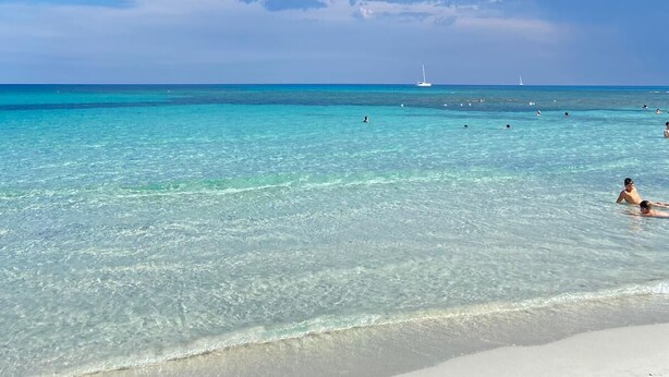 Una playa con agua turquesa cristalina, algunas personas nadando y un velero a lo lejos.