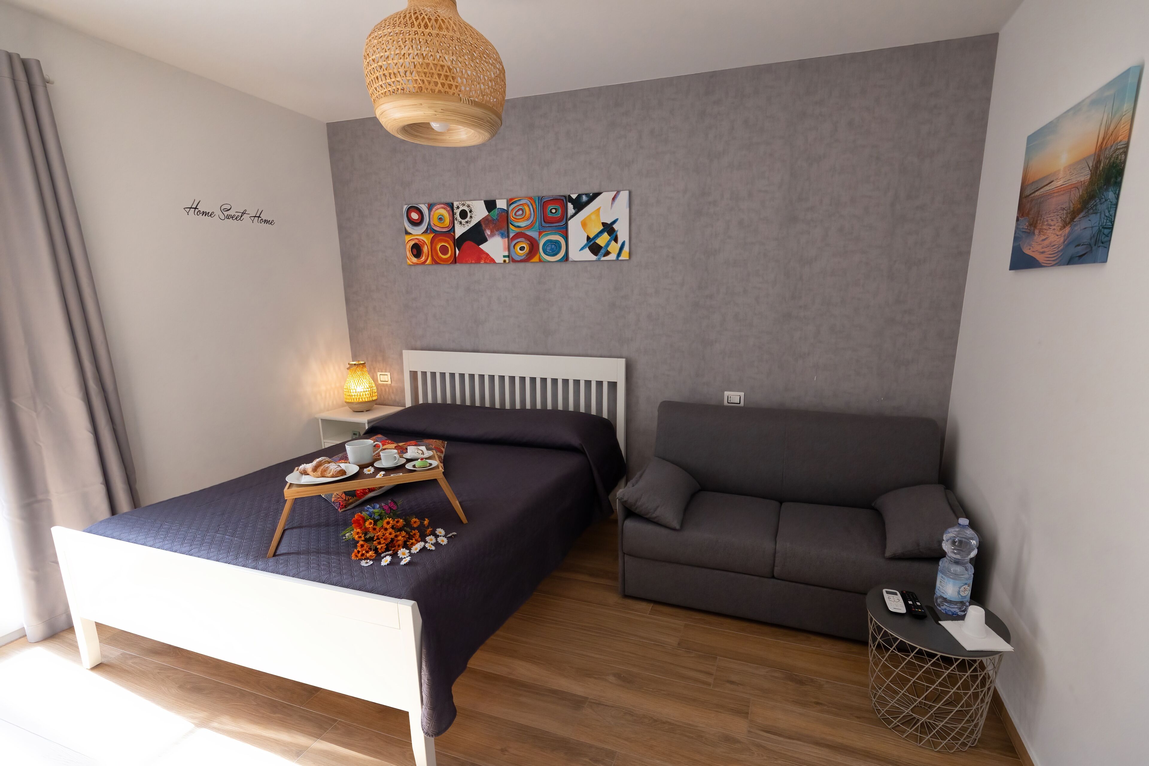 Une chambre à coucher avec un lit, un canapé gris, une petite table d’appoint avec une lampe et un tableau au mur.
