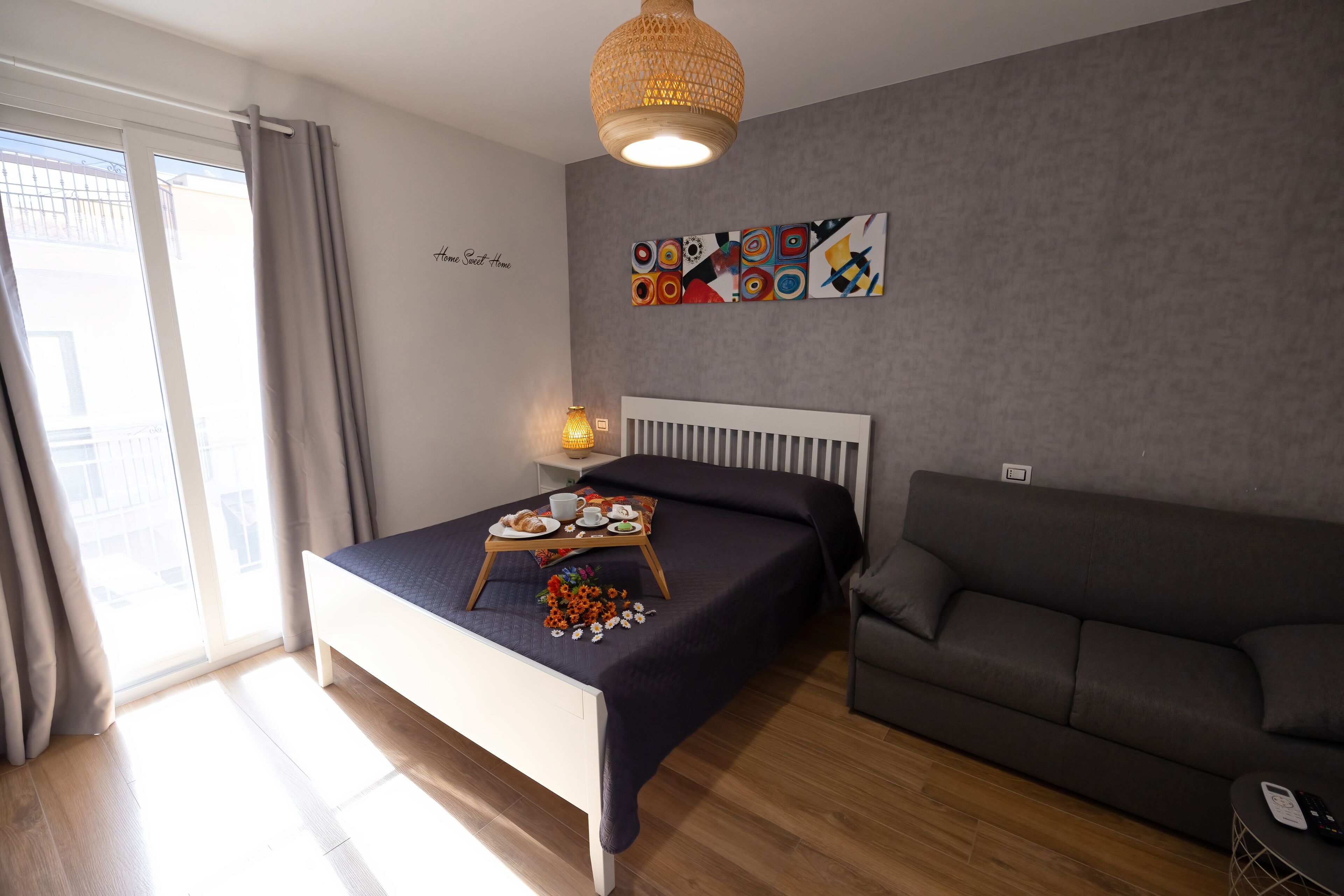 A modern bedroom with a bed, a grey sofa, a bedside table with a lamp, and a wall with abstract art.
