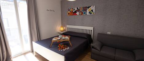 A modern bedroom with a bed, a grey sofa, a bedside table with a lamp, and a wall with abstract art.