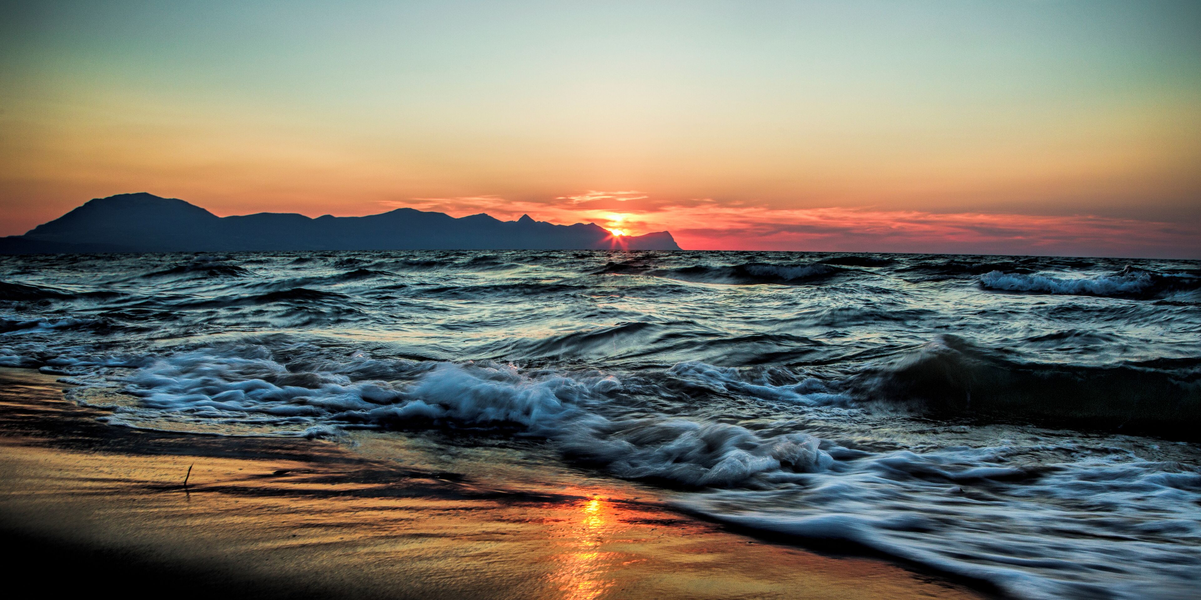 A beach with waves at sunset, mountains in the background, and a clear sky.