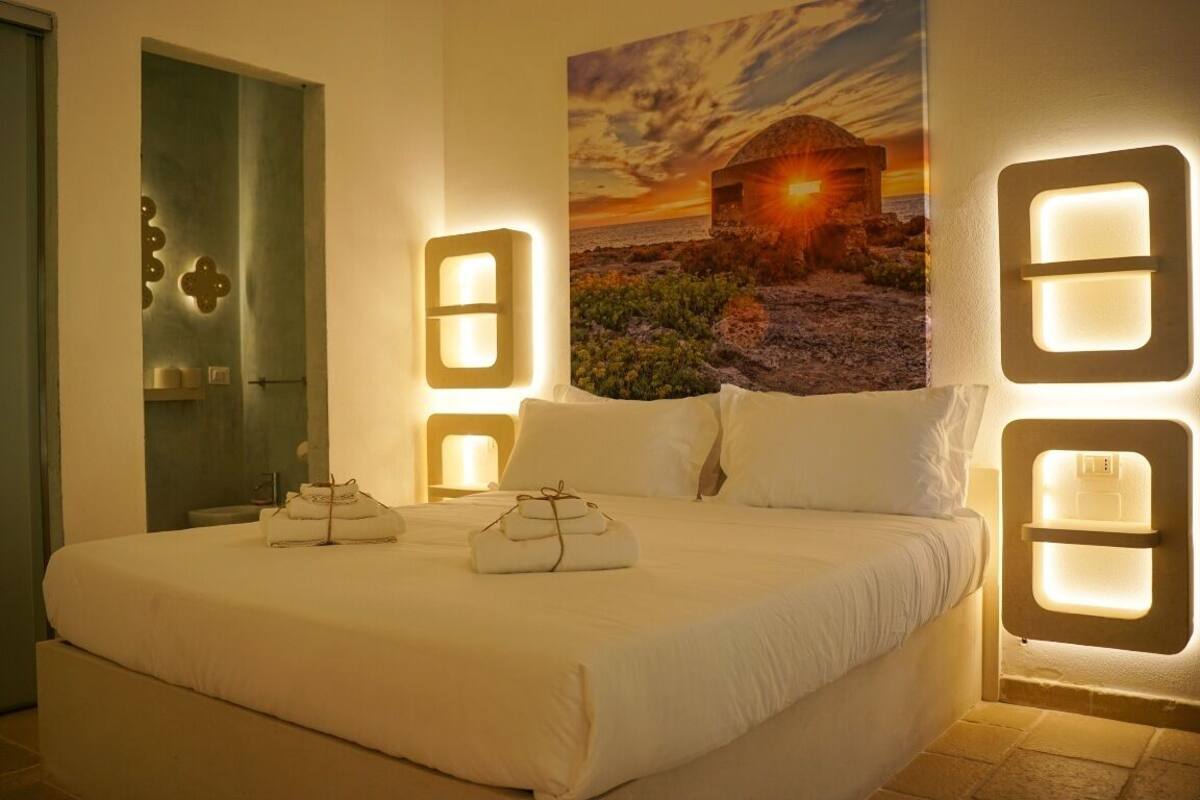 A hotel room with a large bed, a painting of a sunset, and wall-mounted lights.