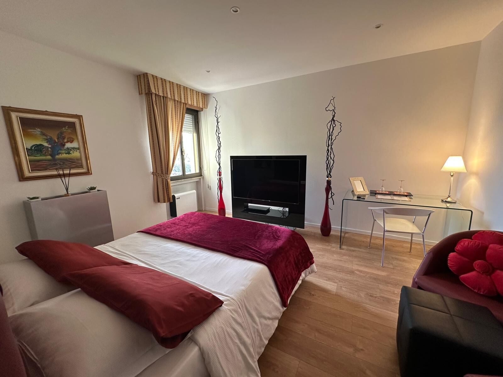 Quarto de hotel com cama, escrivaninha, cadeira, televisão e um quadro na parede.