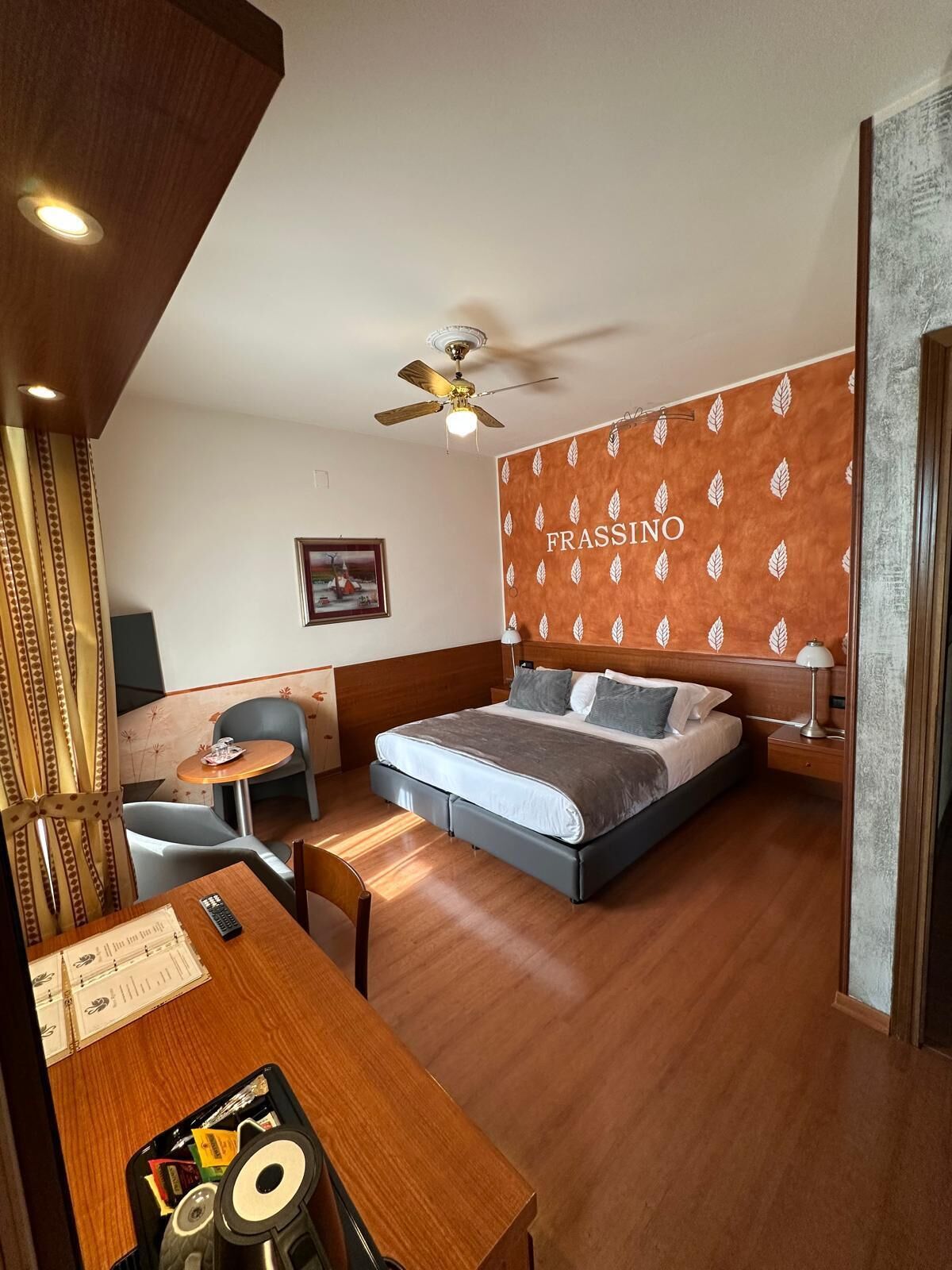 A hotel room with a bed, a desk, a chair, a ceiling fan, and a wall with a leaf pattern.