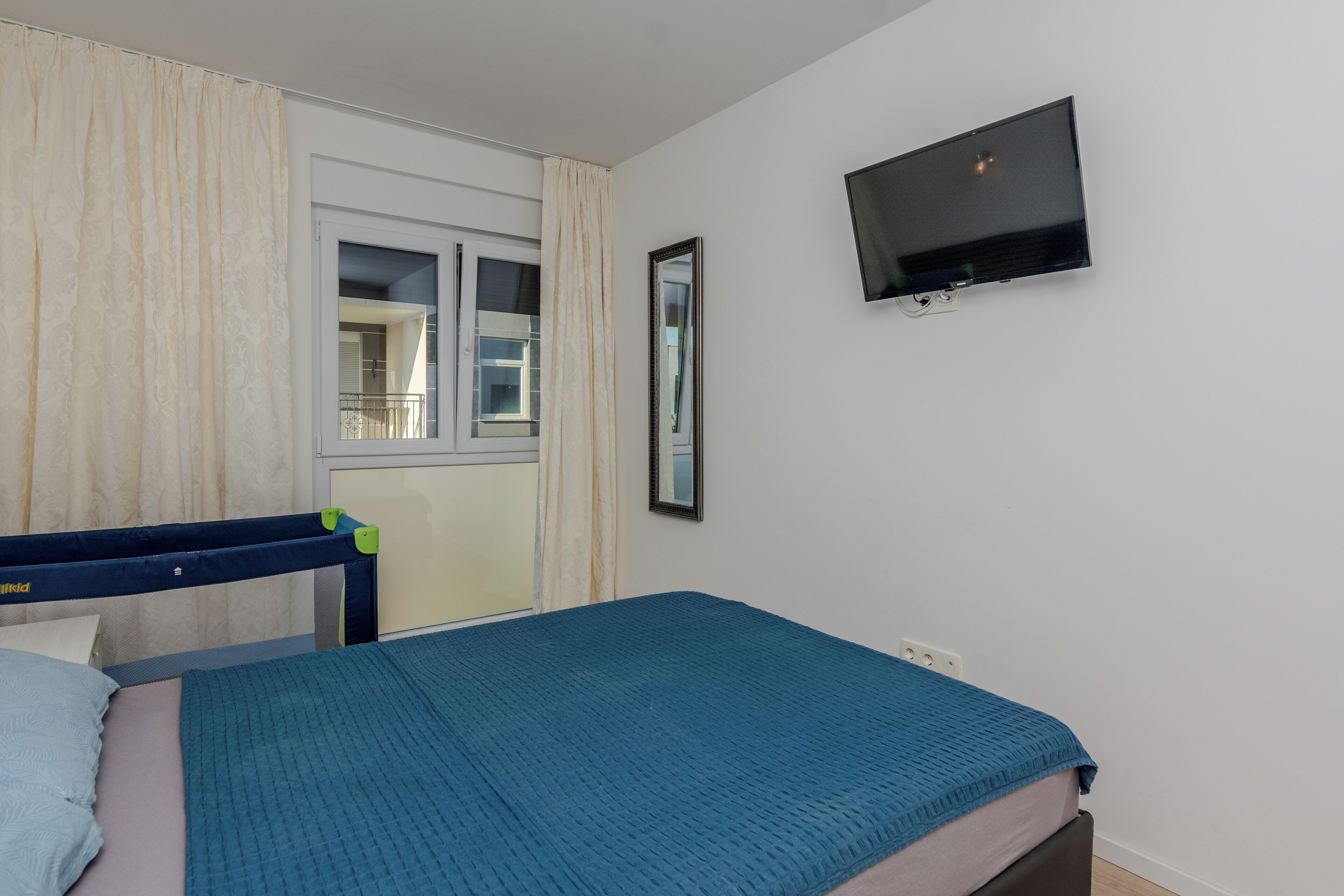 Une chambre avec un lit simple, une fenêtre avec des rideaux, un téléviseur fixé au mur et un miroir.