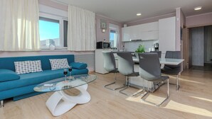 Una sala de estar moderna con un sofá azul, una mesa de centro de cristal y un comedor con una mesa blanca y sillas grises.