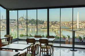 A dining area with large windows offering a view of a cityscape, including a bridge and a mosque. - Csk The Halich Port Istanbul (Istanbul)