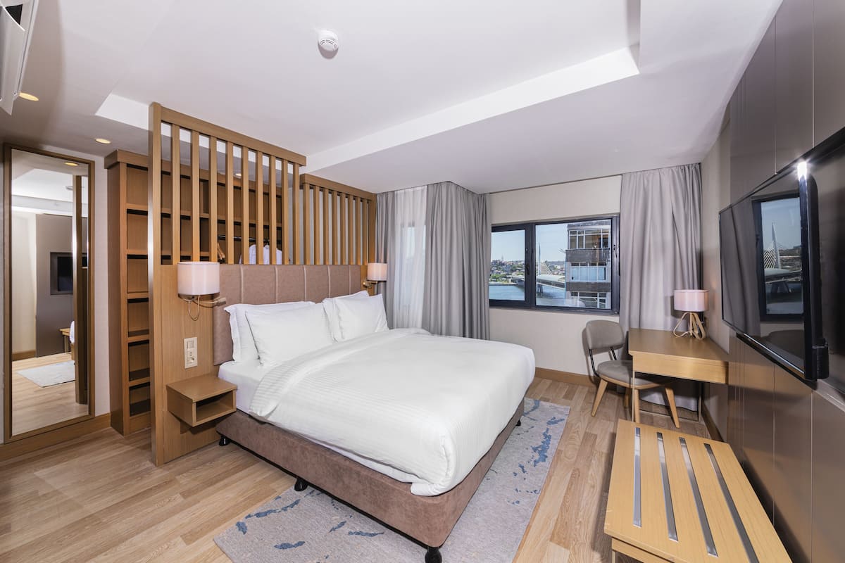 A modern hotel room with a large bed, a desk, a chair, a TV, and a view of the city through the window.