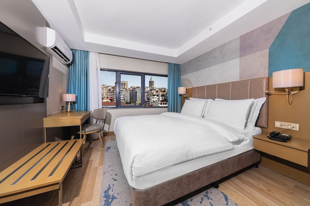 A hotel room with a large bed, a desk, a chair, a TV, and a view of a cityscape.