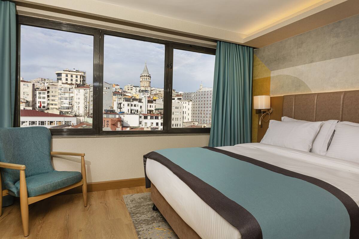 A hotel room with a large bed, a chair, a view of a cityscape through a window, and a lamp on the wall.