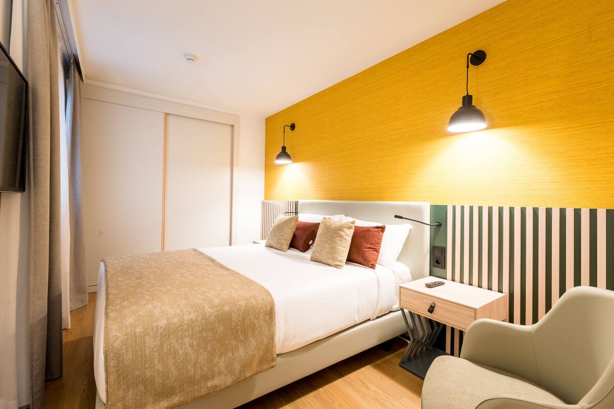 A modern hotel room with a bed, bedside table, chair, and a wooden accent wall.