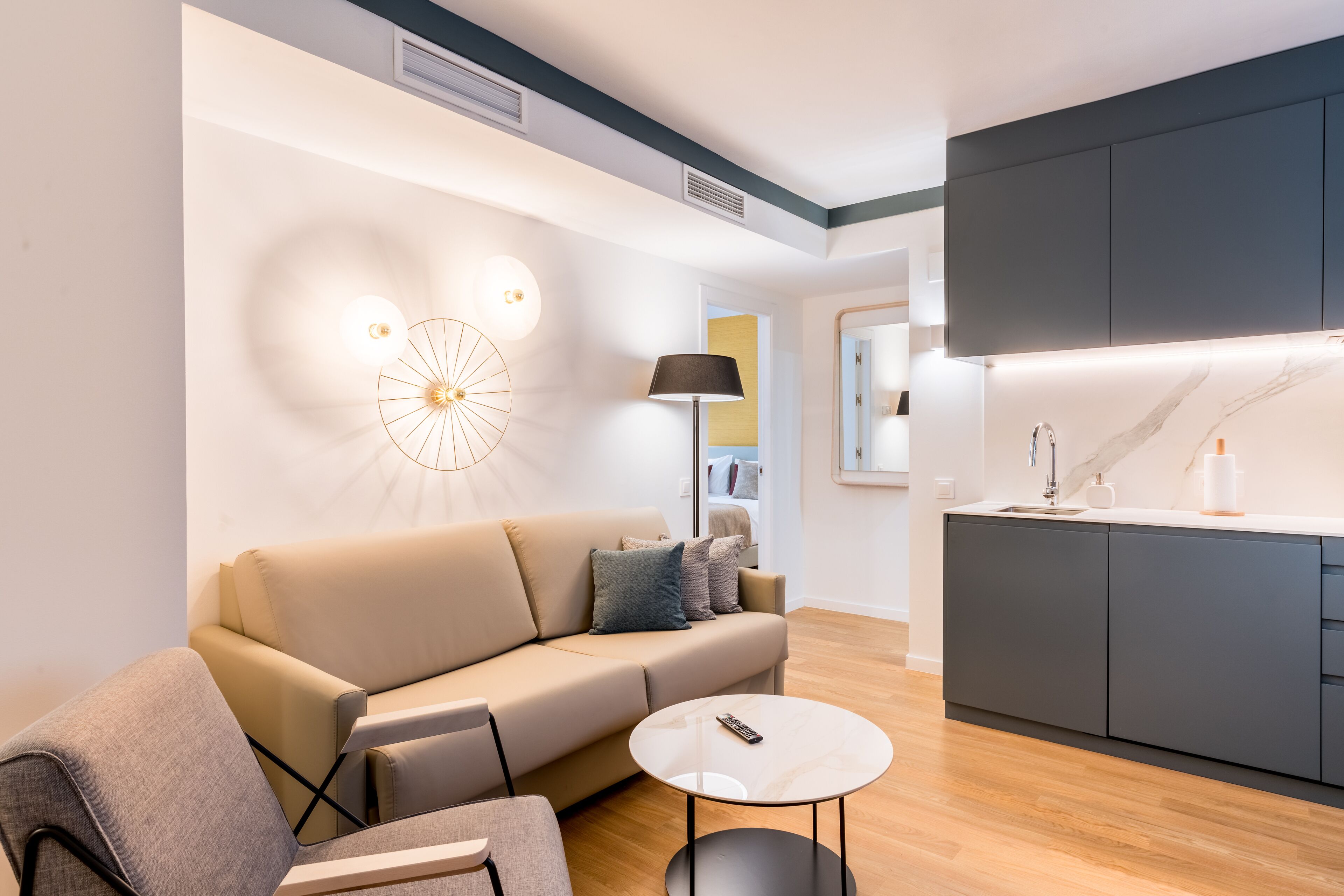 A modern living room with a sofa, a coffee table, and a lamp. There is a kitchen area with cabinets and a countertop.