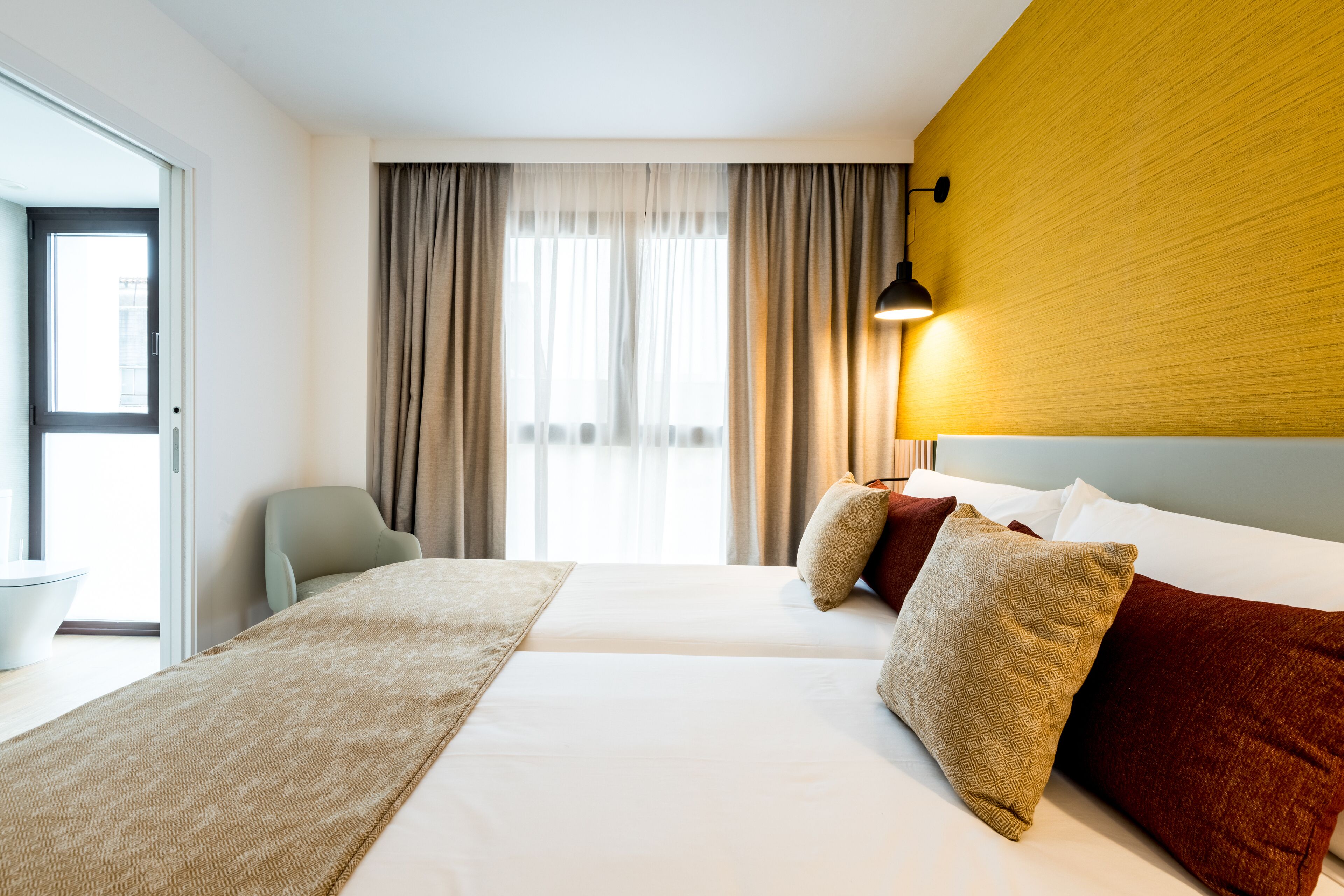 A hotel room with a bed, a chair, a desk, and a window with curtains.