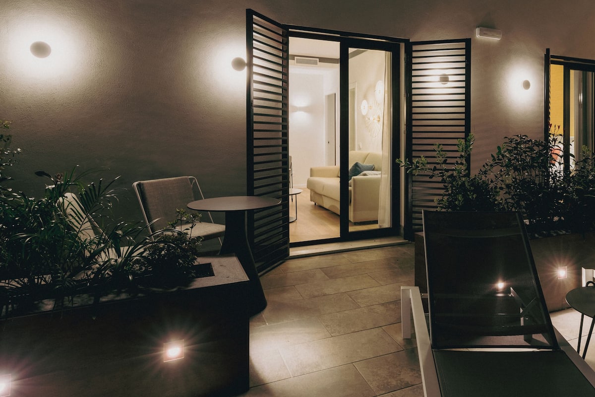 A modern outdoor seating area with chairs and tables, surrounded by potted plants and a sliding glass door leading to an interior space.