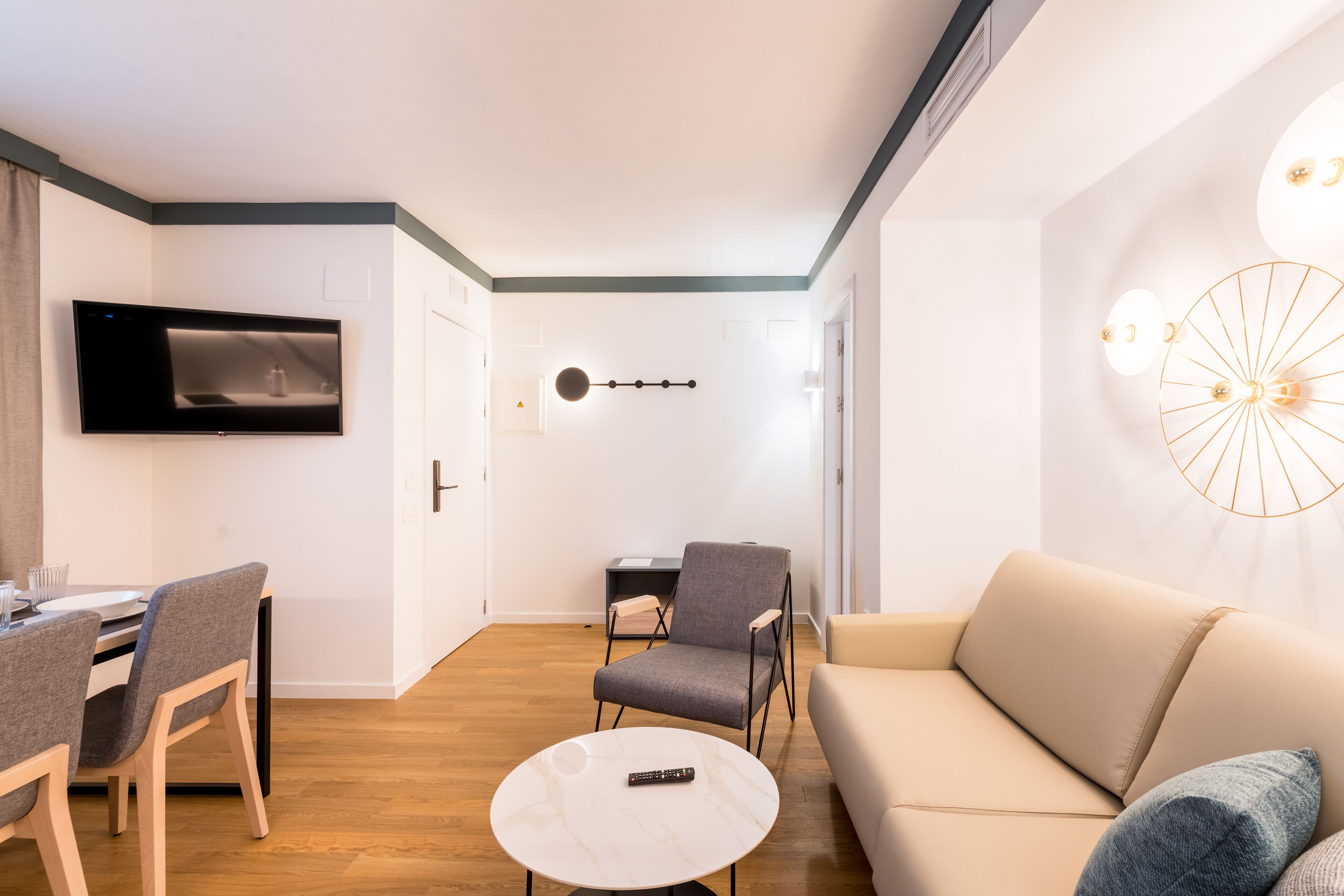 A modern living room with a sofa, a dining table with chairs, a TV mounted on the wall, and a decorative light fixture.