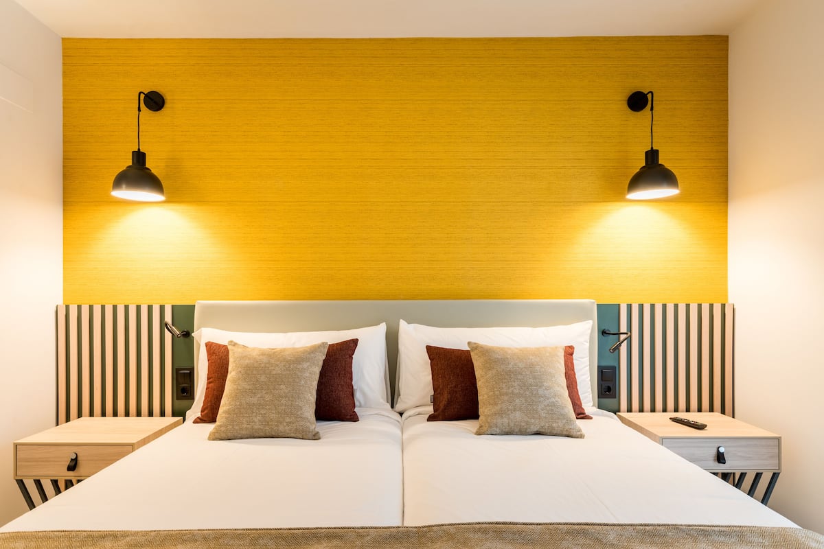 A double bed with two bedside tables, each with a lamp. The headboard has vertical stripes, and the wall above is a solid yellow color.