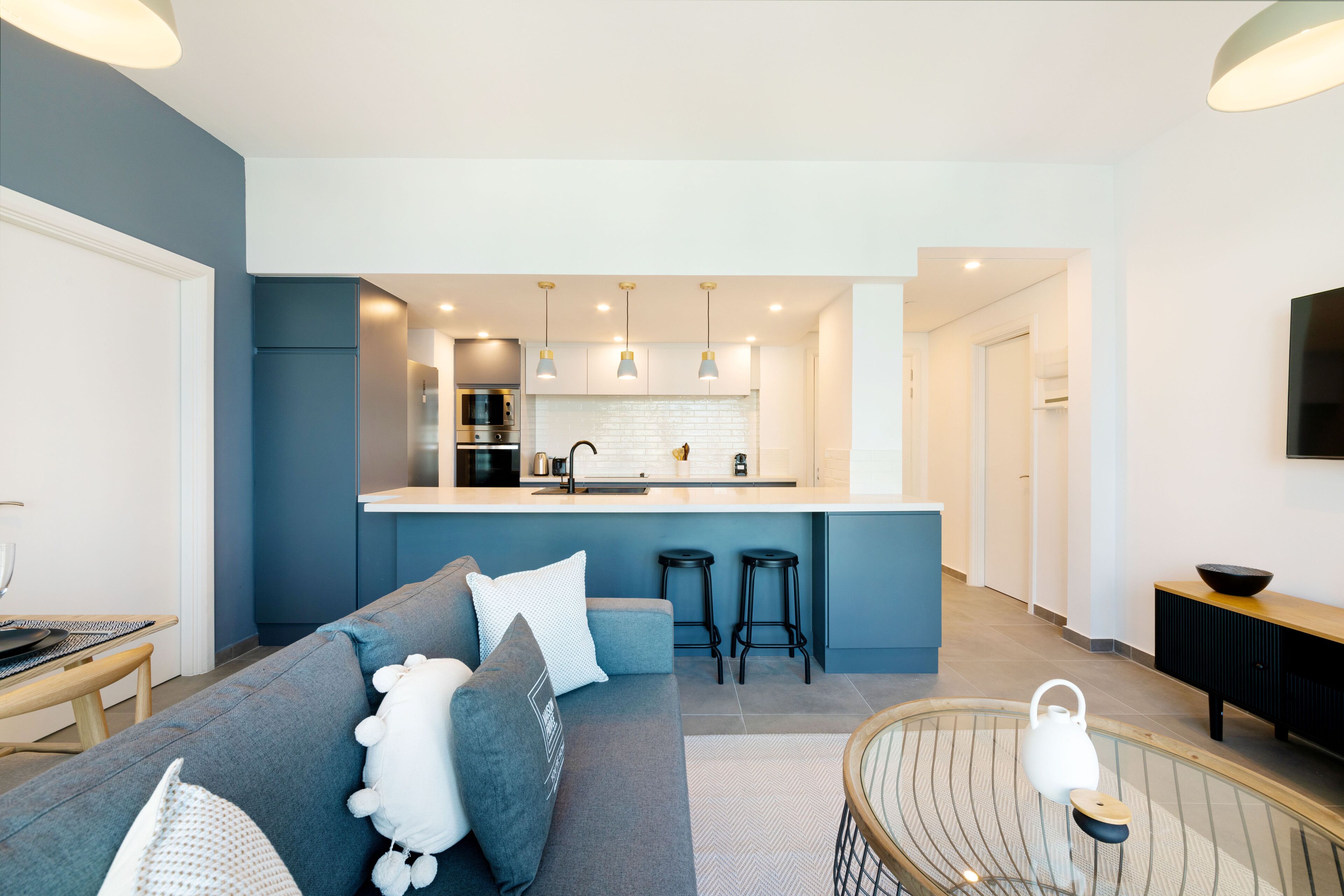 A modern living room with a grey sofa, a coffee table, and a kitchen area with a white countertop and bar stools.