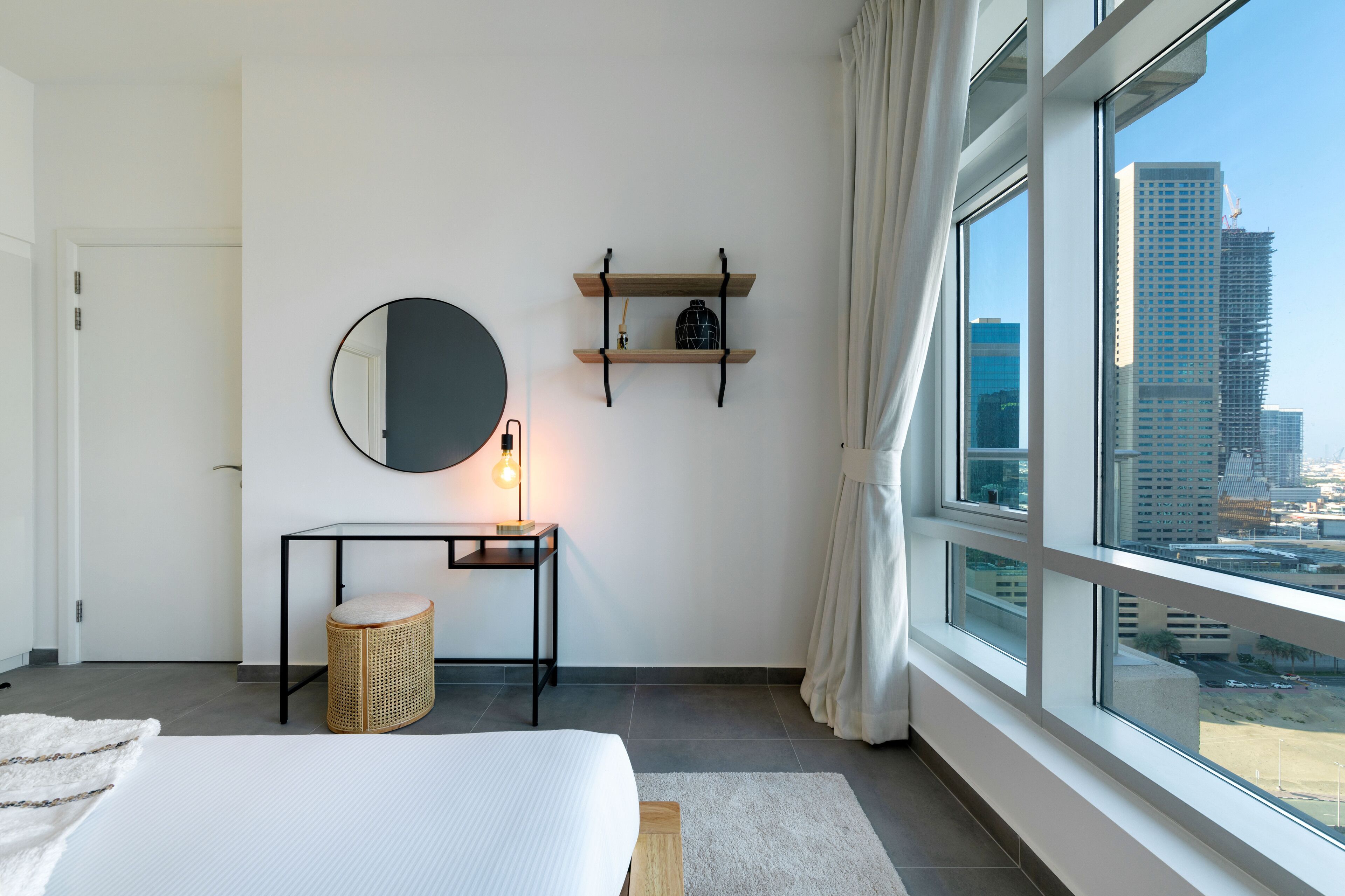 A modern hotel room with a large bed, a round mirror, a small table with a lamp, a wicker stool, and a view of the cityscape through large windows.