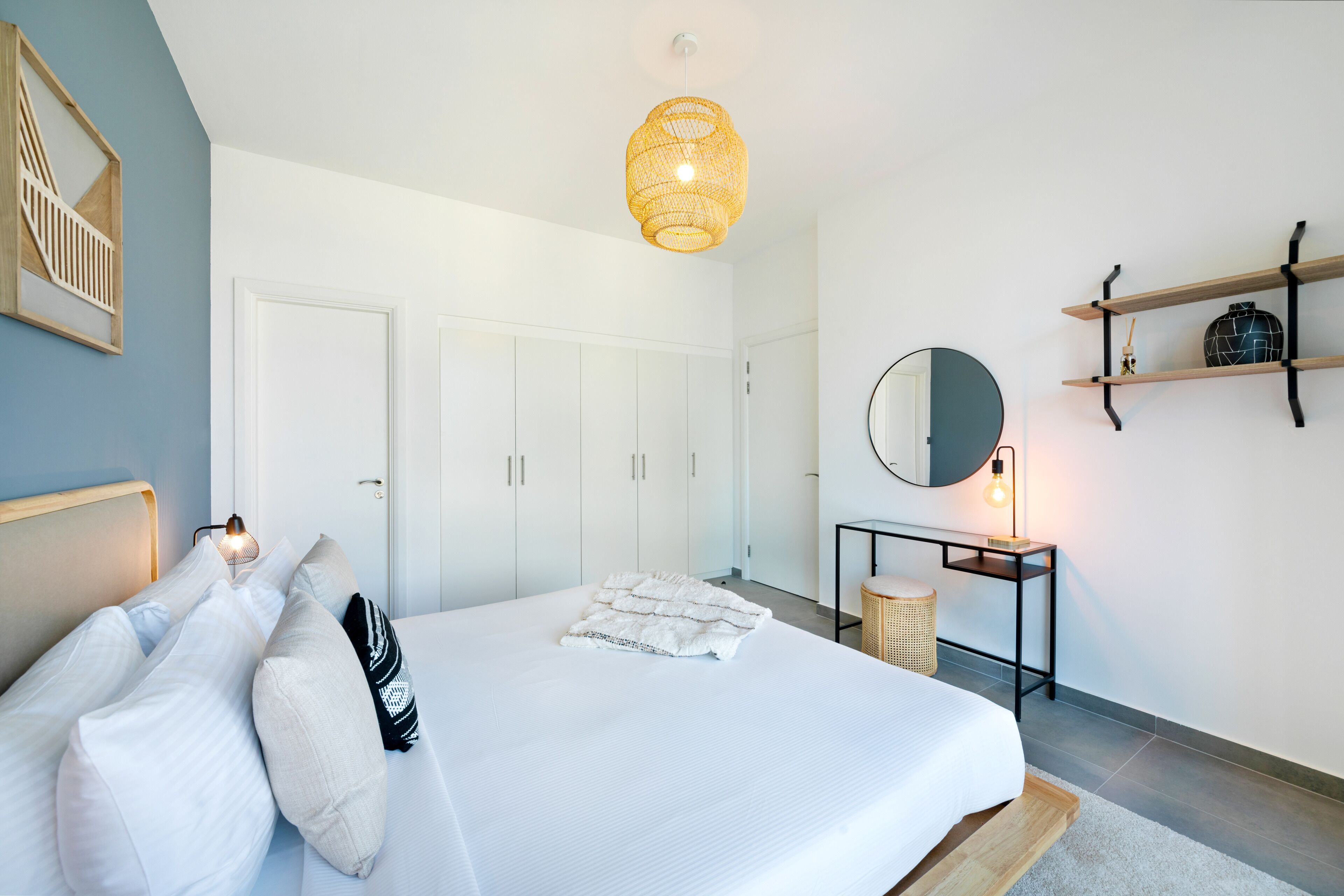 A bedroom with a bed, white bedding, a bedside table with a lamp, a wall-mounted mirror, and a hanging light fixture.