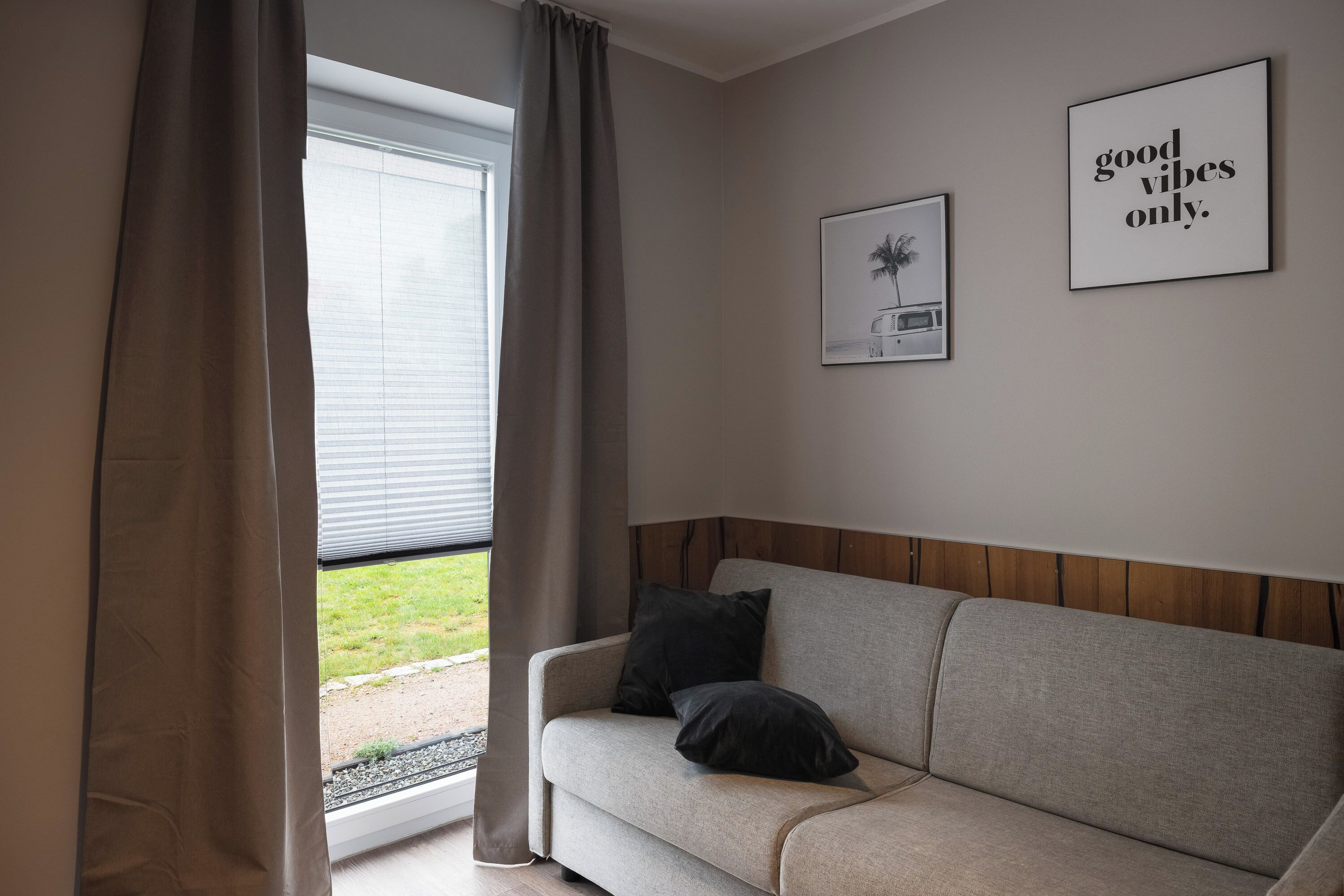 Ein sofas in neutraler Farbe mit zwei schwarzen Kissen, ein Fenster mit Vorhängen und zwei gerahmte Bilder an der Wand.