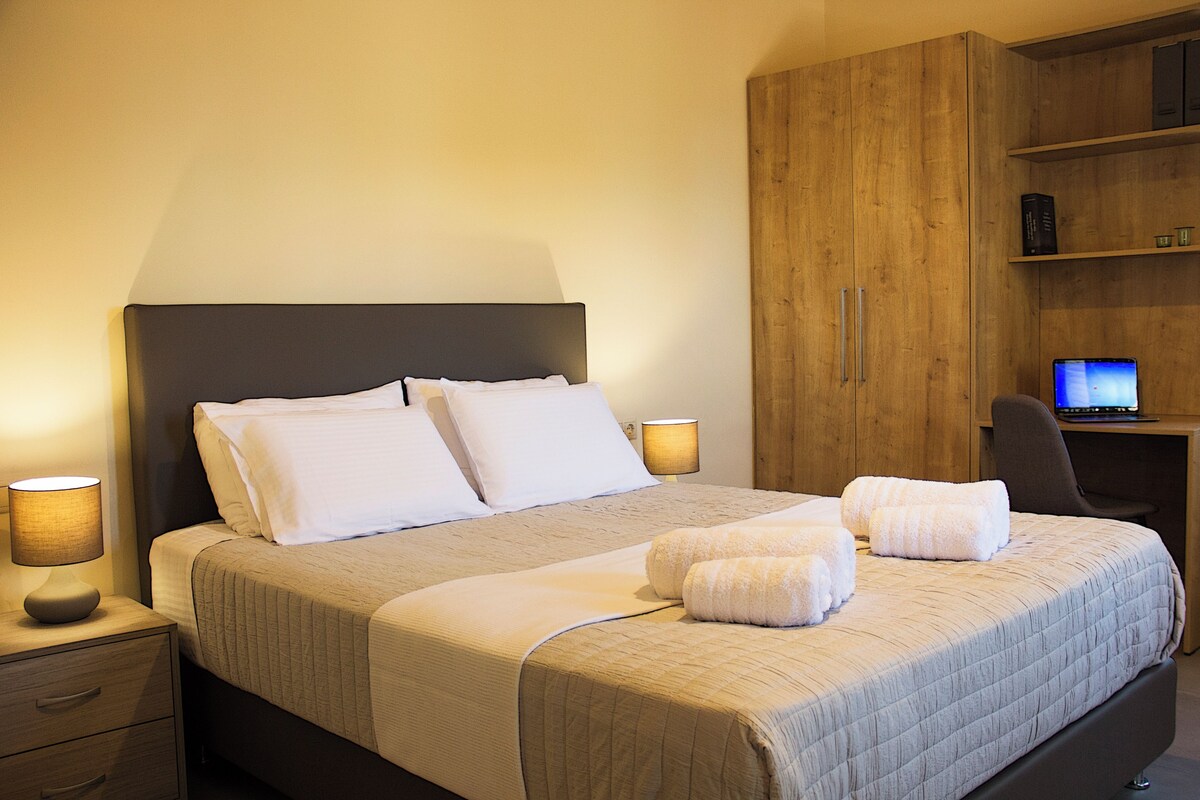 A neatly made bed with pillows and a blanket, a bedside lamp, a wooden wardrobe, and a desk with a computer.