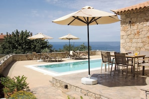 A poolside area with lounge chairs and umbrellas, a stone wall, and a view of the sea.
