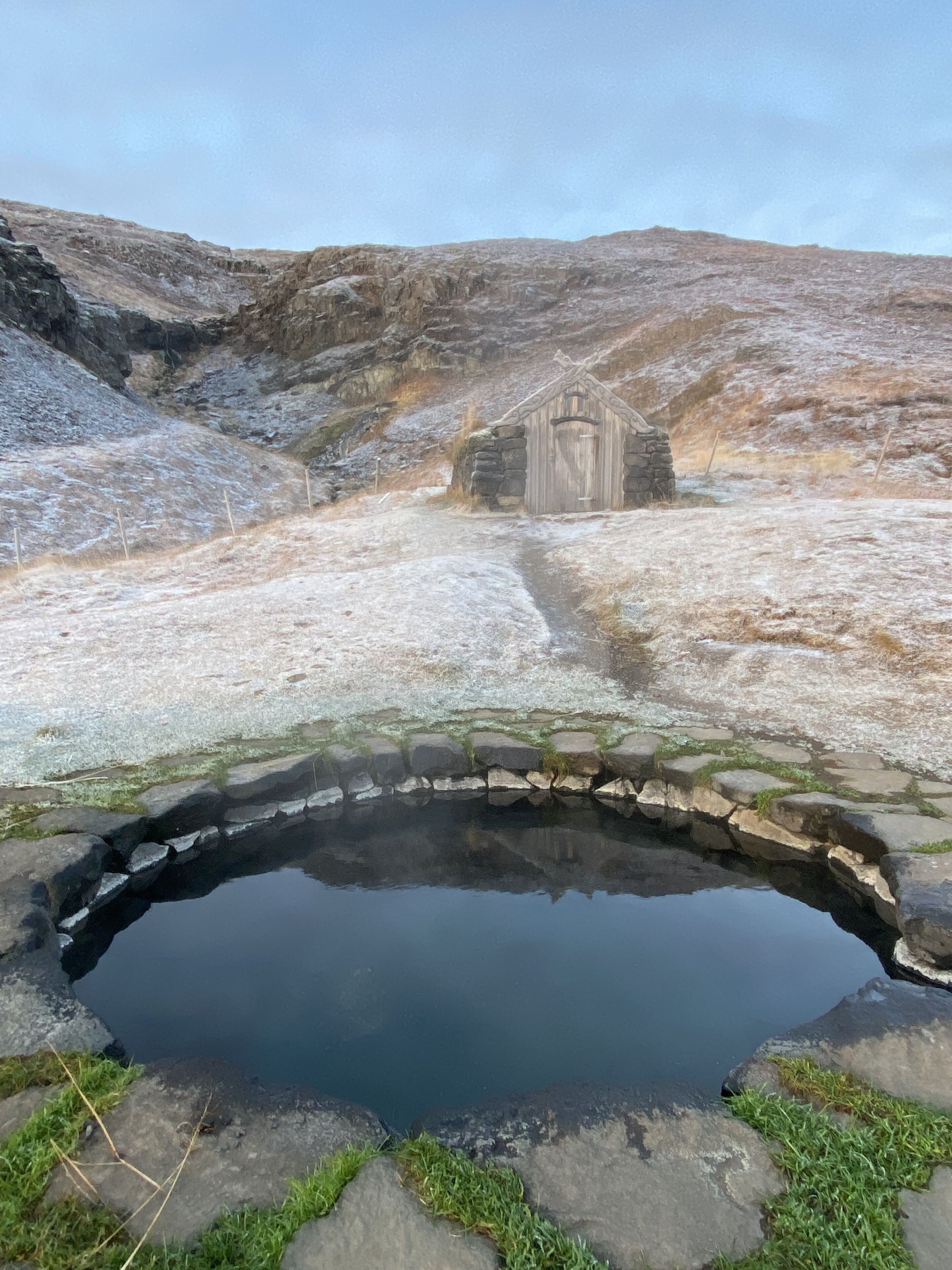 A stone bridge over a reflective water body, leading to a rustic stone building in a hilly landscape.