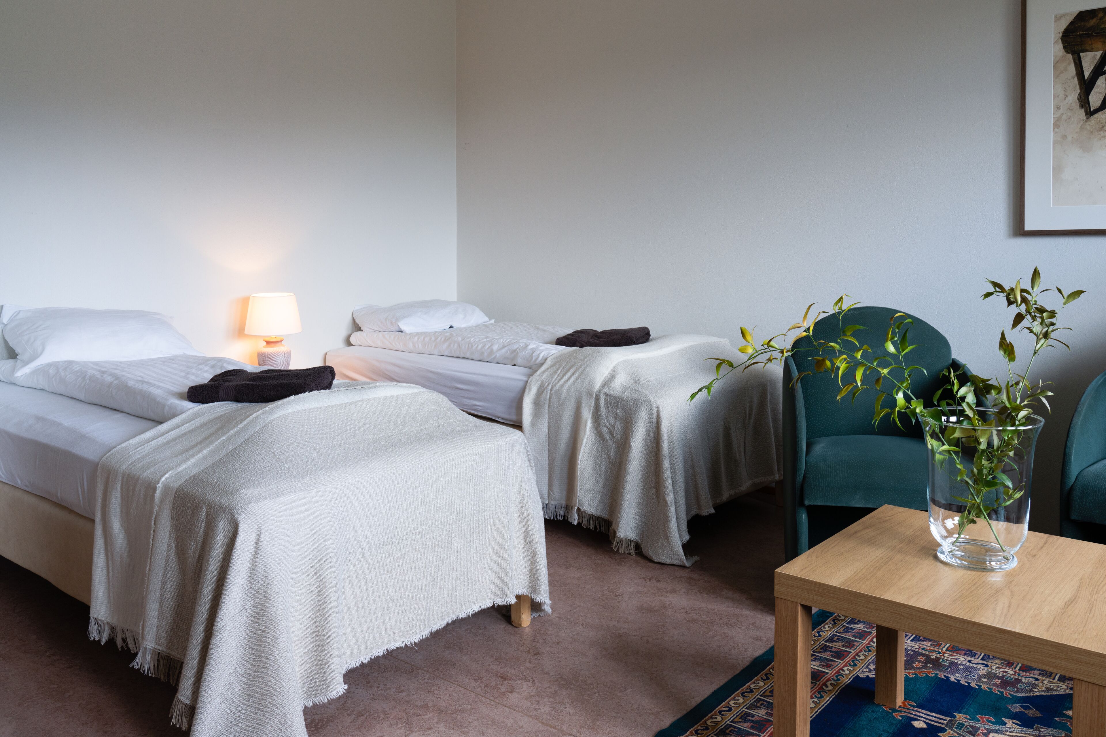 A hotel room with two single beds, a bedside table with a lamp, a chair, and a vase with a plant.