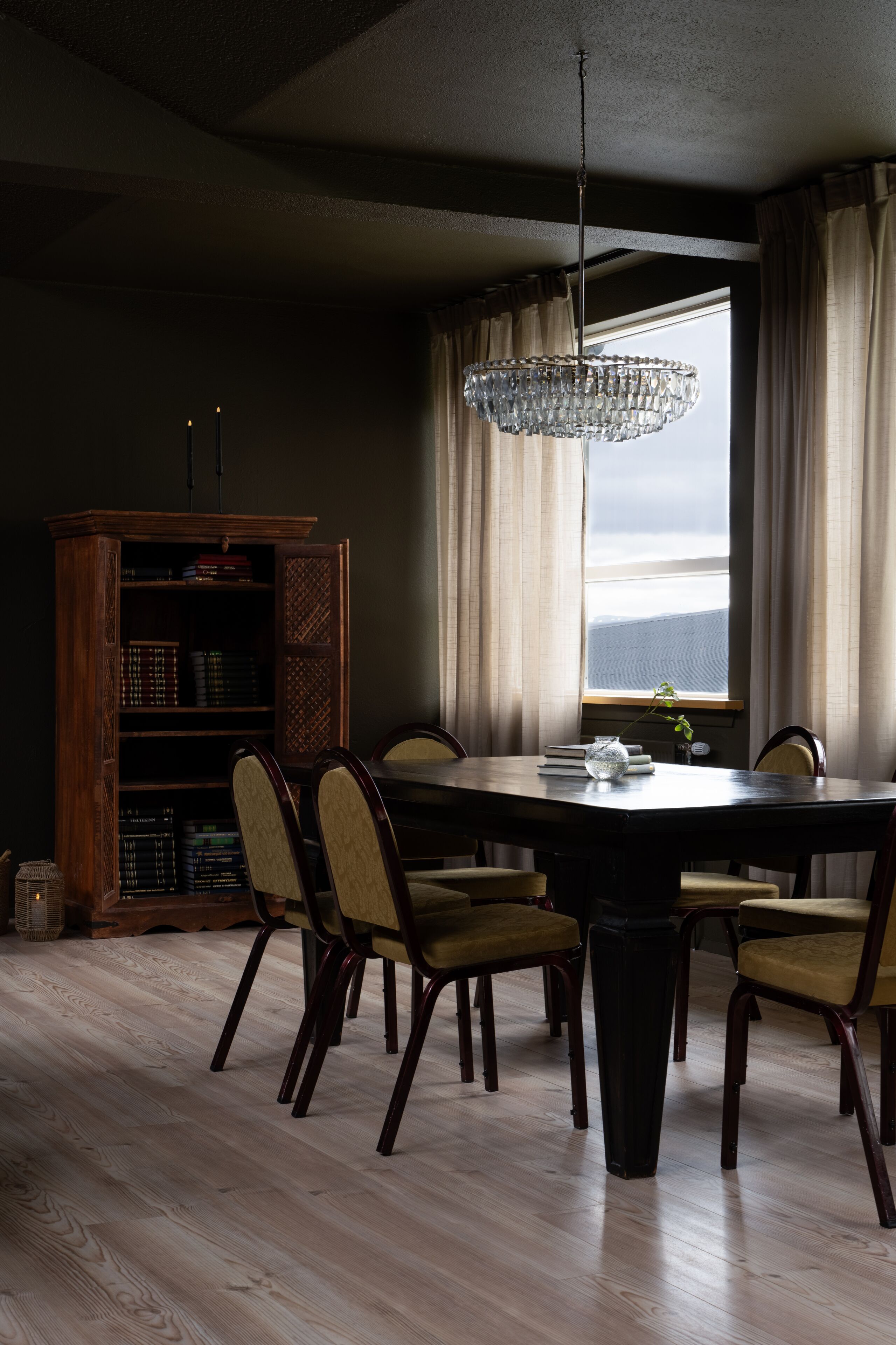 A dining room with a wooden table, six chairs, a chandelier, a bookshelf, and a window with curtains.