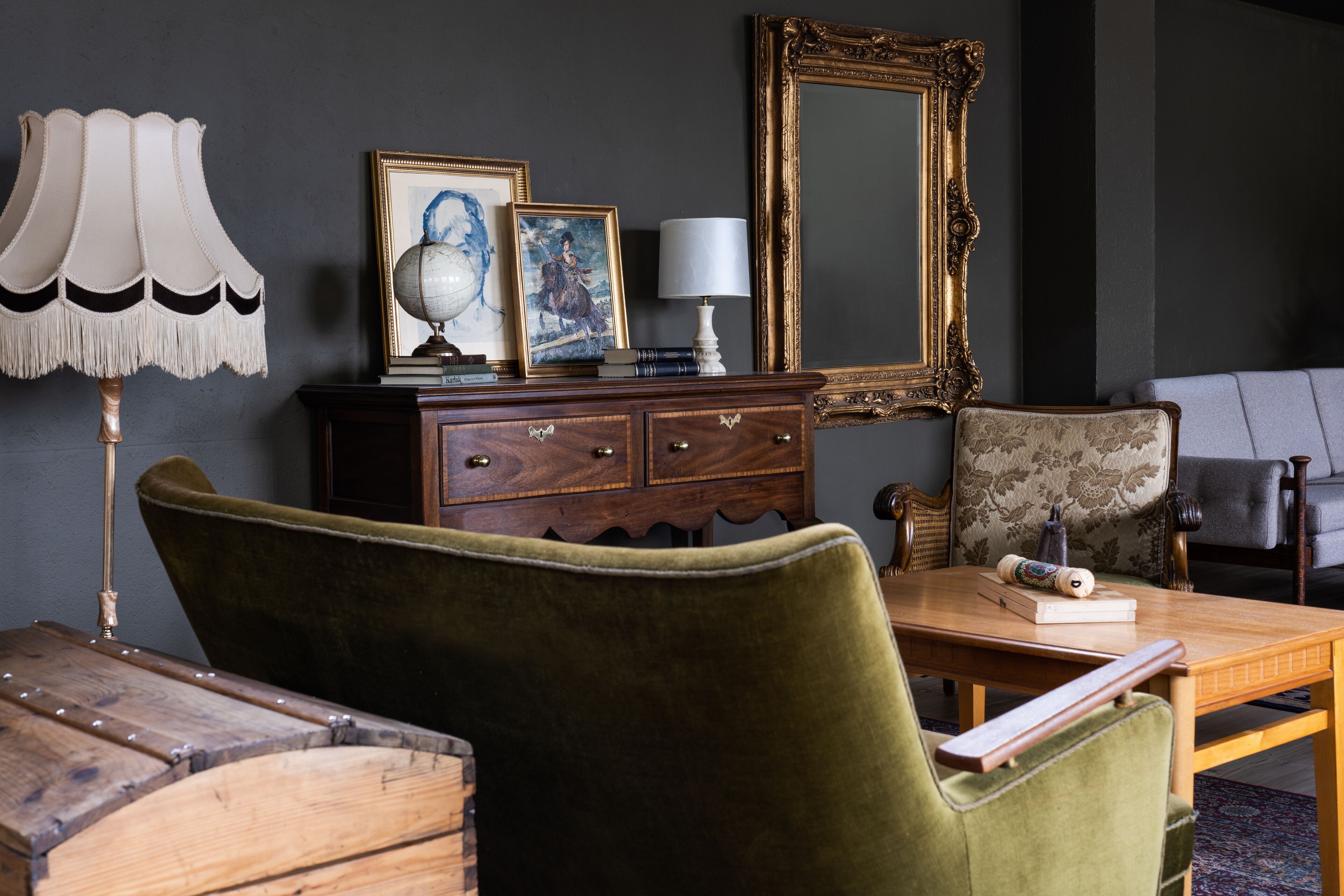 A room with a wooden table, a green upholstered chair, a vintage lamp, a globe, framed artwork, a mirror, and a dark-colored wall.