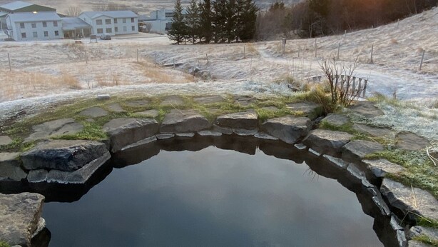 A natural hot spring surrounded by a stone edge, with a backdrop of a sunset over a hilly landscape and a small building in the distance. - Dalahótel (Budardalur)