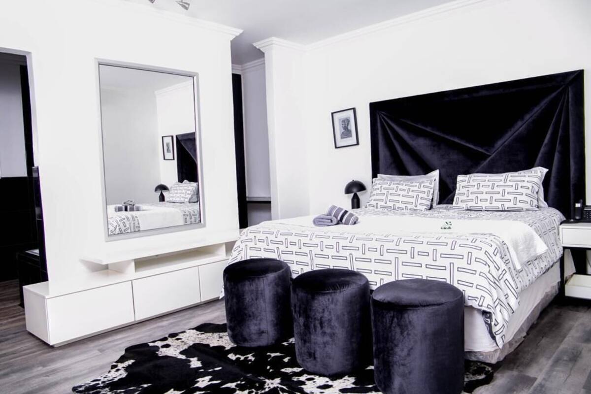 Een moderne slaapkamer met een grote spiegel, een zwart bekleed hoofdeinde, een bed met een gestreept sprei en een kussen van koeienhuid.