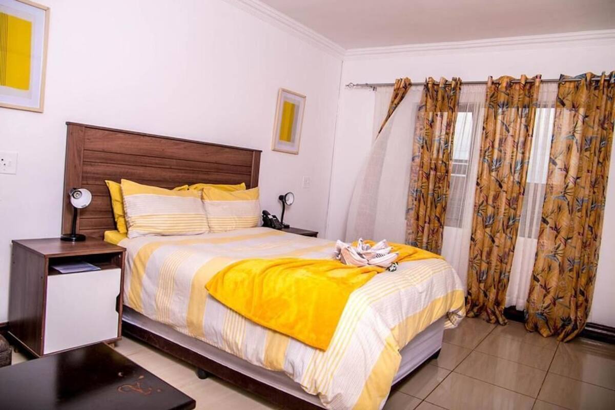 Een netjes opgemaakte slaapkamer met een houten hoofdbord, een bed met geel-witte beddengoed, een nachtkastje met een lamp en een raam met behang.