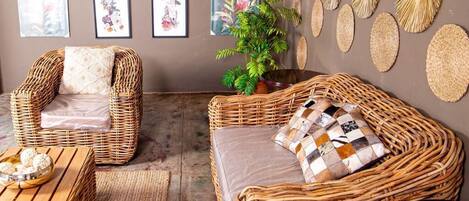 A room with wicker furniture, including two chairs and a sofa, a small table with a bowl of snacks, and a potted plant.