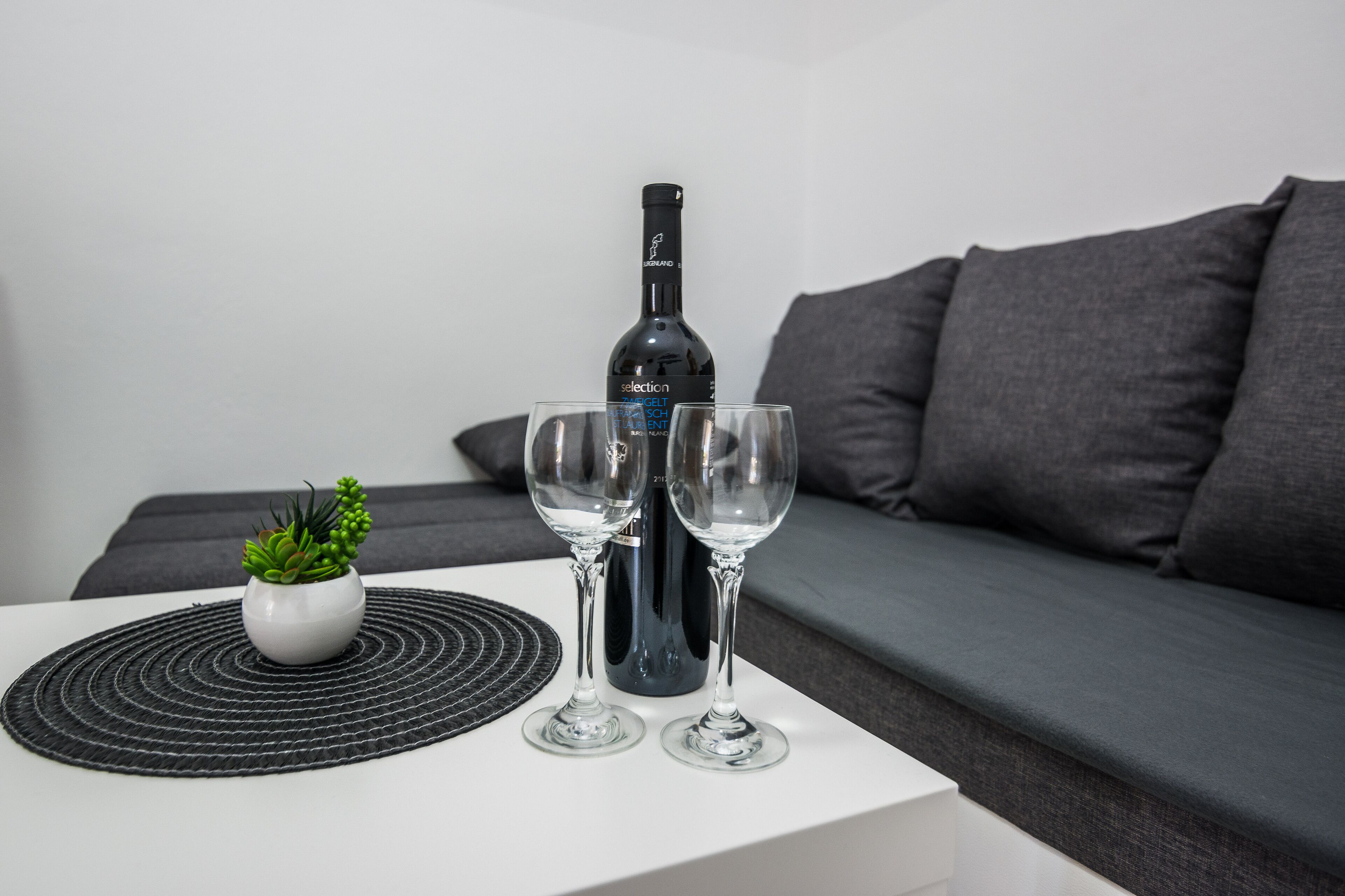 A bottle of wine, two glasses, and a small plant on a white table with a black placemat.