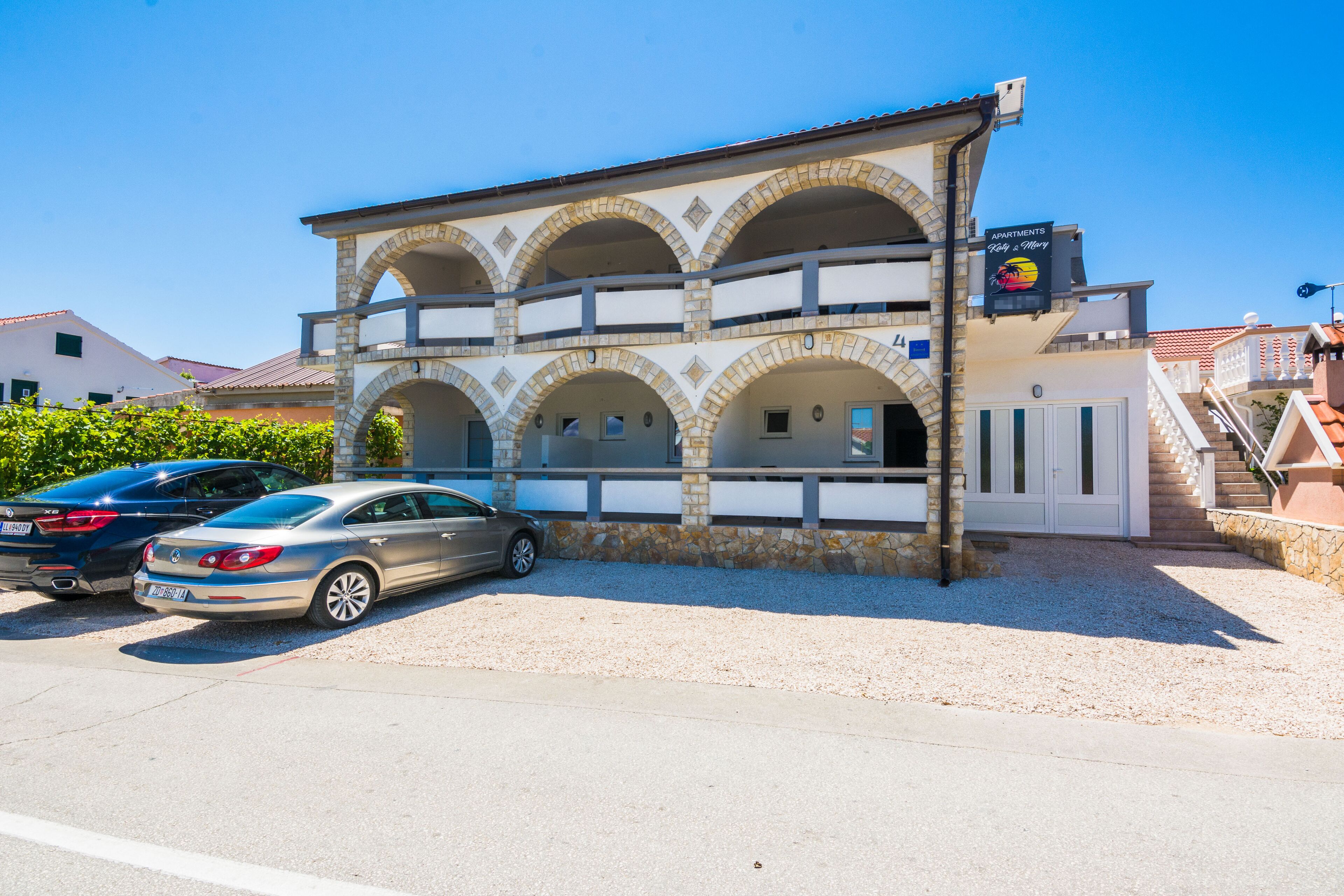 A two-story building with a stone facade and multiple balconies. There are two cars parked in front.