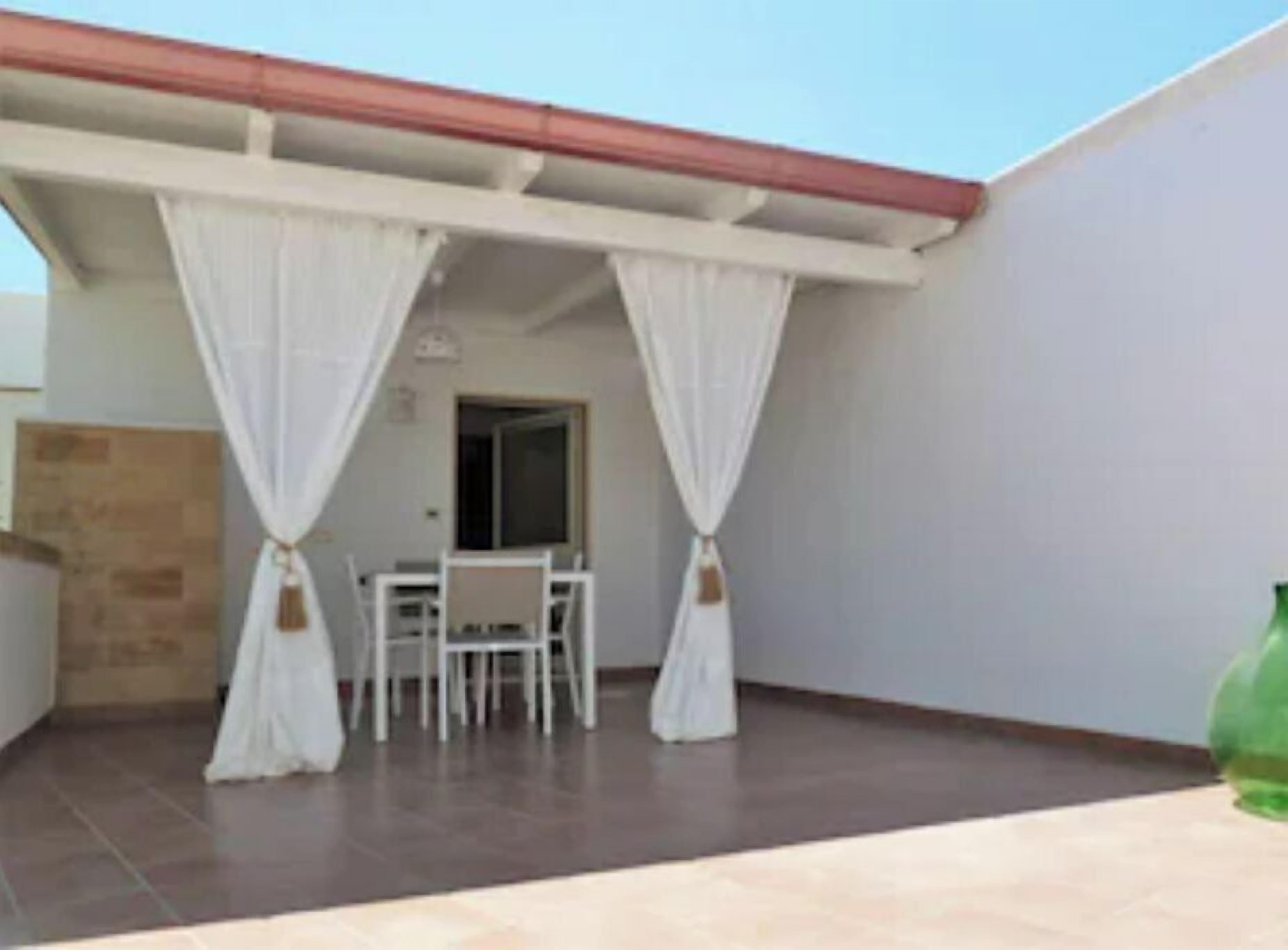 Un porche cubierto con una mesa y sillas, cortinas blancas y una botella verde sobre la mesa.