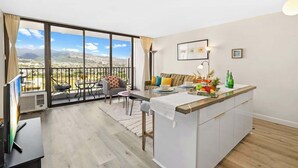 A modern apartment with a balcony, a dining area, a kitchen island, and a large window offering a city view.