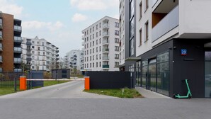 A modern urban street with multi-story residential buildings, a gated entrance, and a green scooter parked by the building. - Stylish Studio | Parking, close to Młociny Metro (Warszawa)