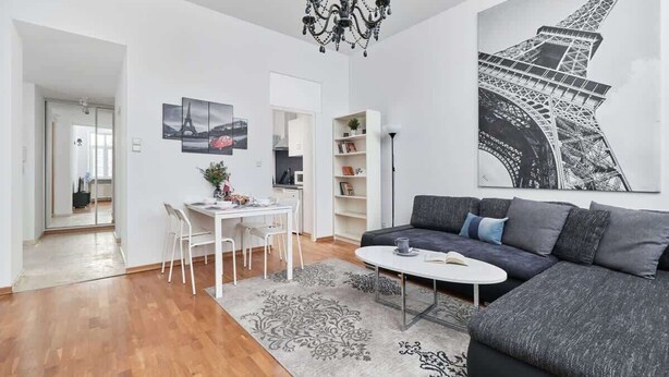A modern living room with a dark grey sofa, a round coffee table, and a chandelier. There is a dining area with a white table and chairs, and a bookshelf with various items. A large black and white Eiffel Tower artwork is on the wall.