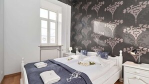 A bedroom with a white bed, dark patterned wallpaper, a window with white blinds, and a white nightstand with a lamp.