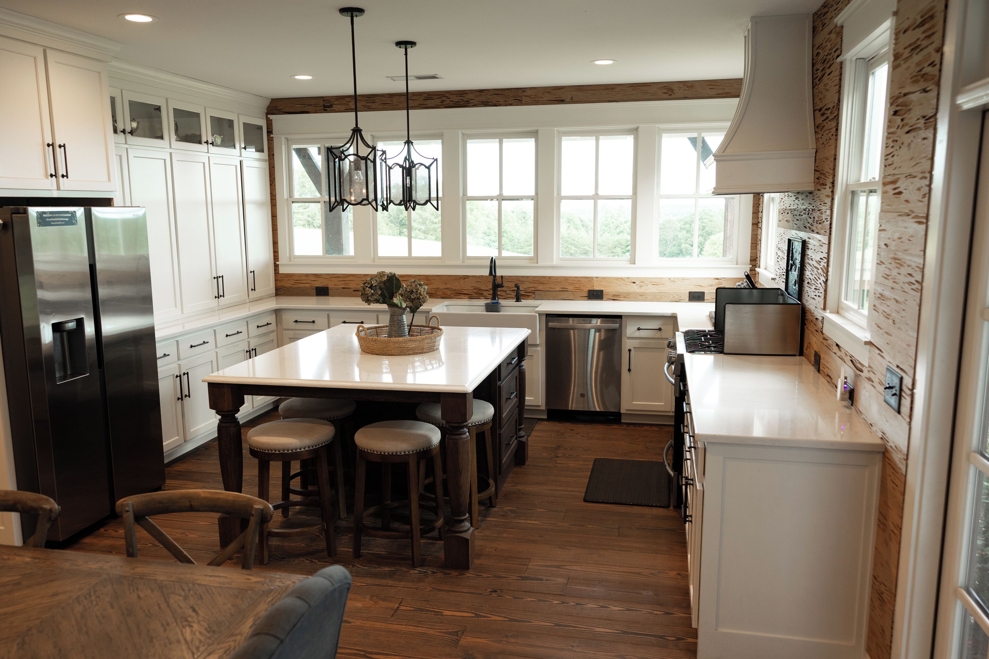 A modern kitchen with white cabinetry, a central island with stools, a stainless steel refrigerator, and a large window providing natural light.