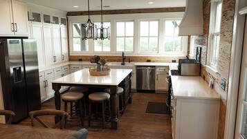 A modern kitchen with white cabinetry, a central island with stools, a stainless steel refrigerator, and a large window providing natural light.